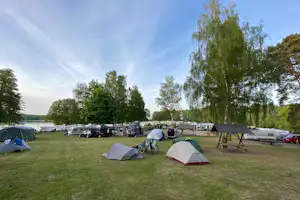 Zeltplatz mit mehreren Zelten und Wohnwagen auf einer Rasenfläche, Tische und Stühle am Rand, Bäume und Himmel im Hintergrund.