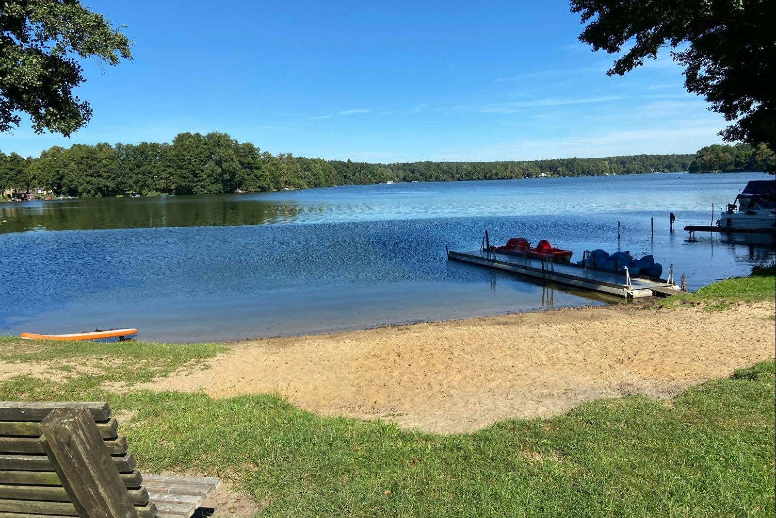 Ruhiger See mit Steg und Booten, sandiger Uferbereich und Rasen am Campingplatz, Bäume und blauer Himmel im Hintergrund.