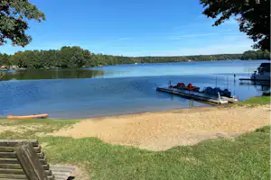 Ruhiger See mit Steg und Booten, sandiger Uferbereich und Rasen am Campingplatz, Bäume und blauer Himmel im Hintergrund.