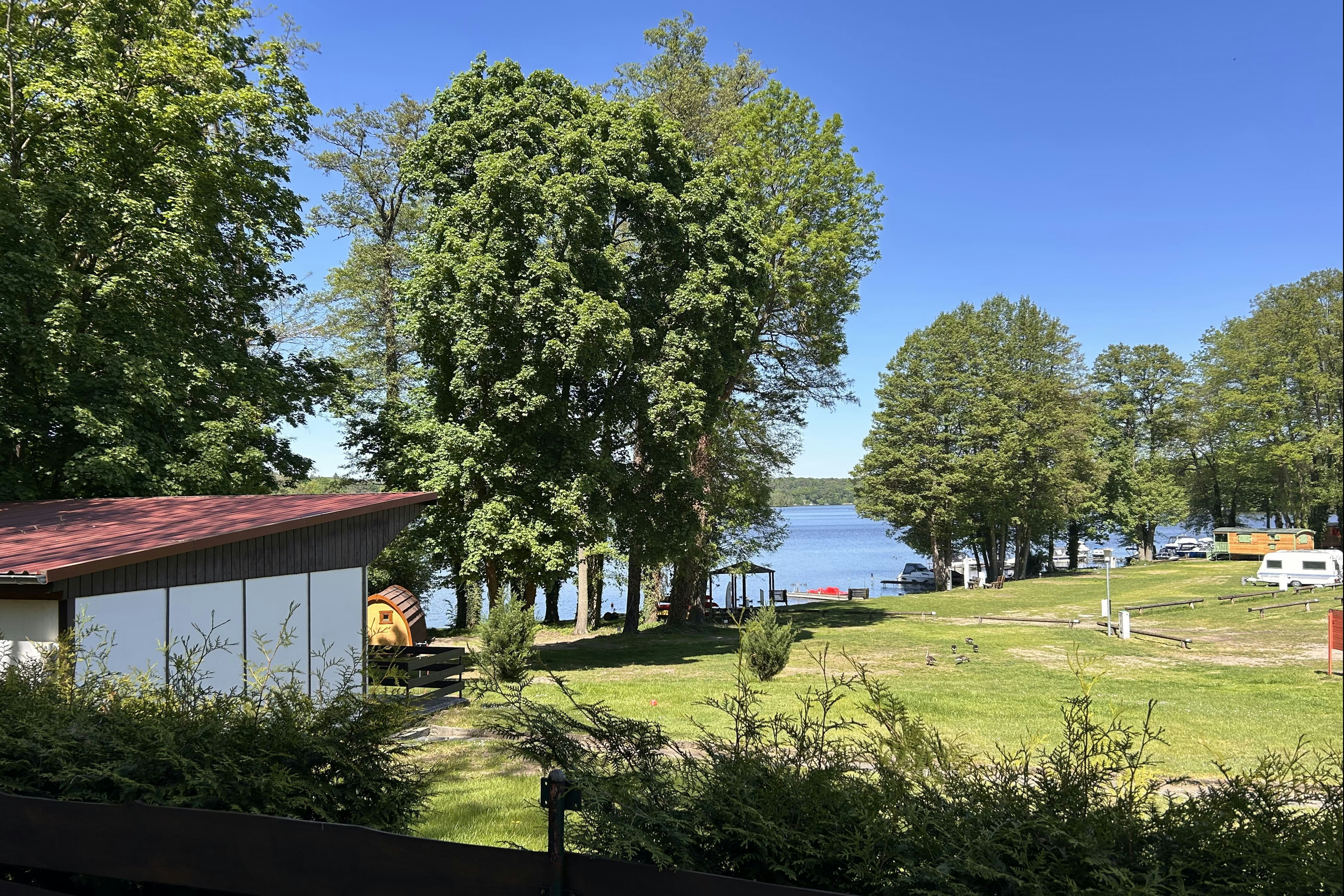 Blick auf einen Campingplatz mit Rasen, Bäumen und einem kleinen Gebäude am Ufer, im Hintergrund Wasser und Wohnwagen.