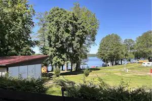 Blick auf einen Campingplatz mit Rasen, Bäumen und einem kleinen Gebäude am Ufer, im Hintergrund Wasser und Wohnwagen.