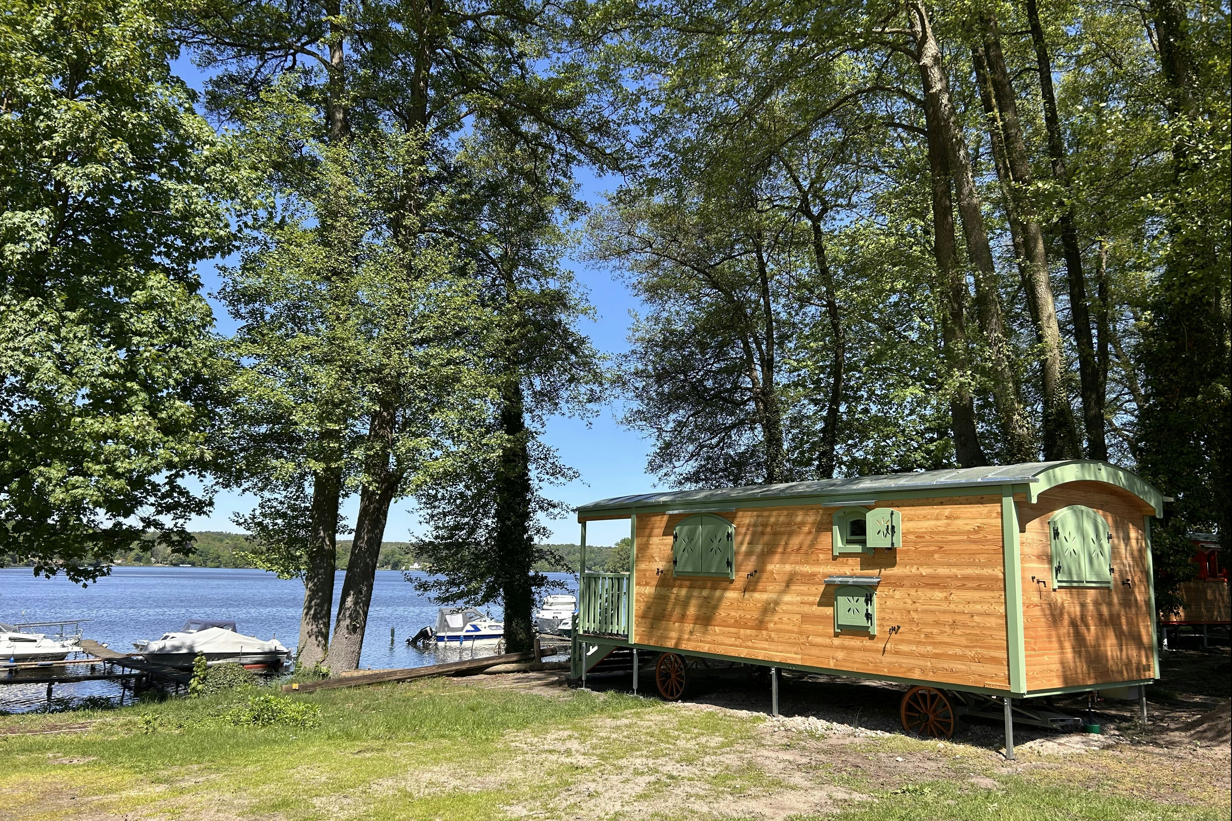 Holz-Blockhaus mit grünen Fenstern und Terrasse am See, umgeben von Bäumen und Rasen, im Hintergrund Boote am Ufer.