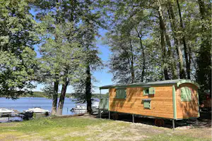Holz-Blockhaus mit grünen Fenstern und Terrasse am See, umgeben von Bäumen und Rasen, im Hintergrund Boote am Ufer.