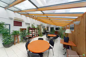 Überdachte Terrasse mit orangefarbenen Tischen, Stühlen, Pflanzenkübeln und Holzverkleidung an der weißen Gebäudewand.