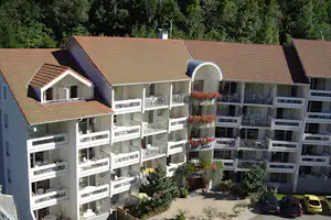 Mehrstöckiges Apartmentgebäude mit Terrassen und Balkonen, weißem Geländer, Ziegeldach und Blumenampeln vor dichtem Grün.