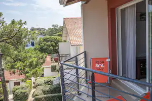 Balkon mit rotem Stuhl und Geländer an einem Wohngebäude, Blick auf Hecken, Bäume und Dächer im Hintergrund.