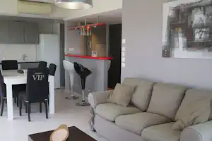 Wohnzimmer mit beigem Sofa, Esstisch und Stühlen, Küchenbereich im Hintergrund, große Fensterfront und Wandbild.