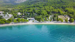 Luftaufnahme eines Campingplatzes am See mit Uferpromenade, Bäumen und Gebäuden am Hang.