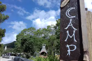 Holzschild am Wegesrand auf einem Campingplatz, dahinter Bäume, Hecken und ein schmaler Weg zwischen Stellplätzen.