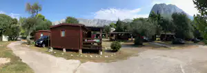 Holzhütte und Stellplätze auf einem Campingplatz, mit Auto, Bäumen und Blick auf Berge im Hintergrund.