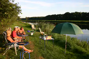Camping am See: Zelt mit Sonnenschirm, Campingstühle und Tisch am Ufer, im Hintergrund Bäume und Wasser.