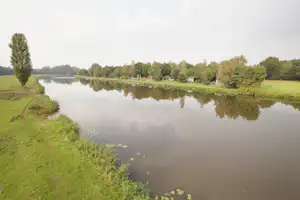 Ruhiger See am Campingplatz mit Grasufer, Bäumen und kleinen Gebäuden am Rand, gespiegelt im Wasser.