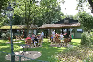 Sitzgruppe im Campinggarten mit Stühlen und Tischen auf dem Rasen, neben Holzhäusern und unter Bäumen.