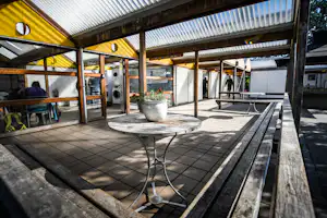 Überdachte Camping-Terrasse mit Grillbereich, Tisch und Sitzgelegenheiten, Ziegelboden und Holzgeländer, im Hintergrund Gebäude und Pflanzen.