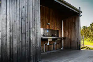 Holz-Campinghaus außen mit überdachter Terrasse und Küchenzeile mit Spüle, daneben ein Weg und Grünfläche.