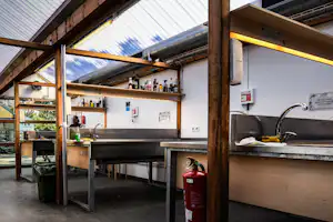 Überdachter Küchenbereich im Campingplatz: Spülbecken, Arbeitsflächen, Regale und ein Feuerlöscher an der Wand.