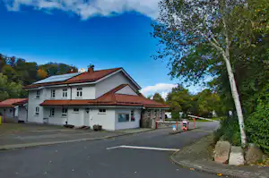 Weißes Campinghaus mit rotem Ziegeldach, Parkplatz und Gehweg, daneben ein Baum und Hecke am Rand.
