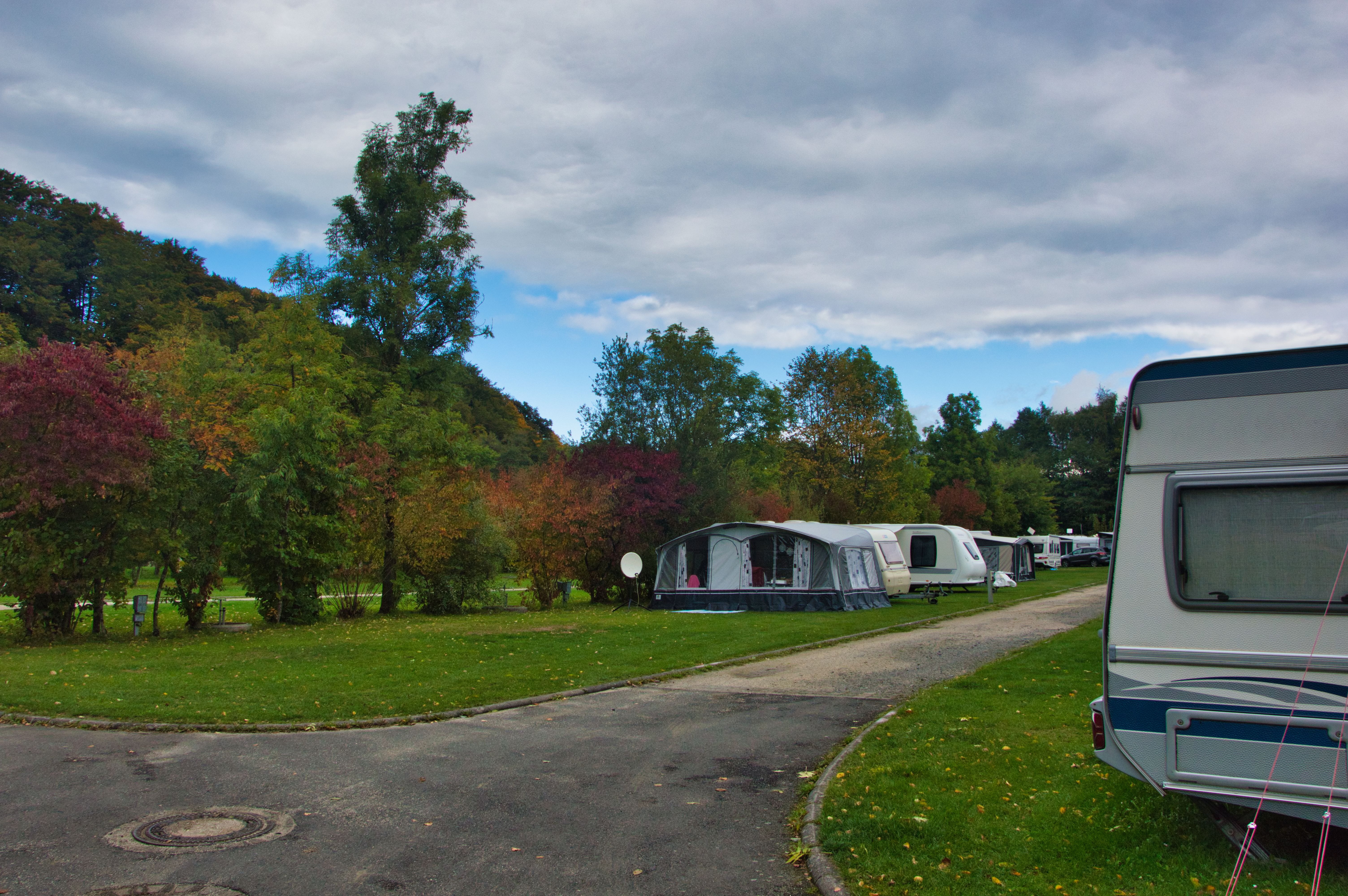 Wohnwagen auf einem Campingplatz-Stellplatz neben einer Zufahrtsstraße, mit Rasenfläche und Bäumen im Hintergrund.