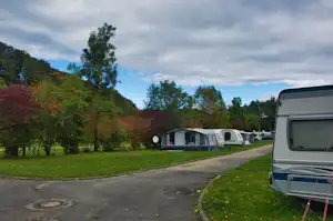Wohnwagen auf einem Campingplatz-Stellplatz neben einer Zufahrtsstraße, mit Rasenfläche und Bäumen im Hintergrund.