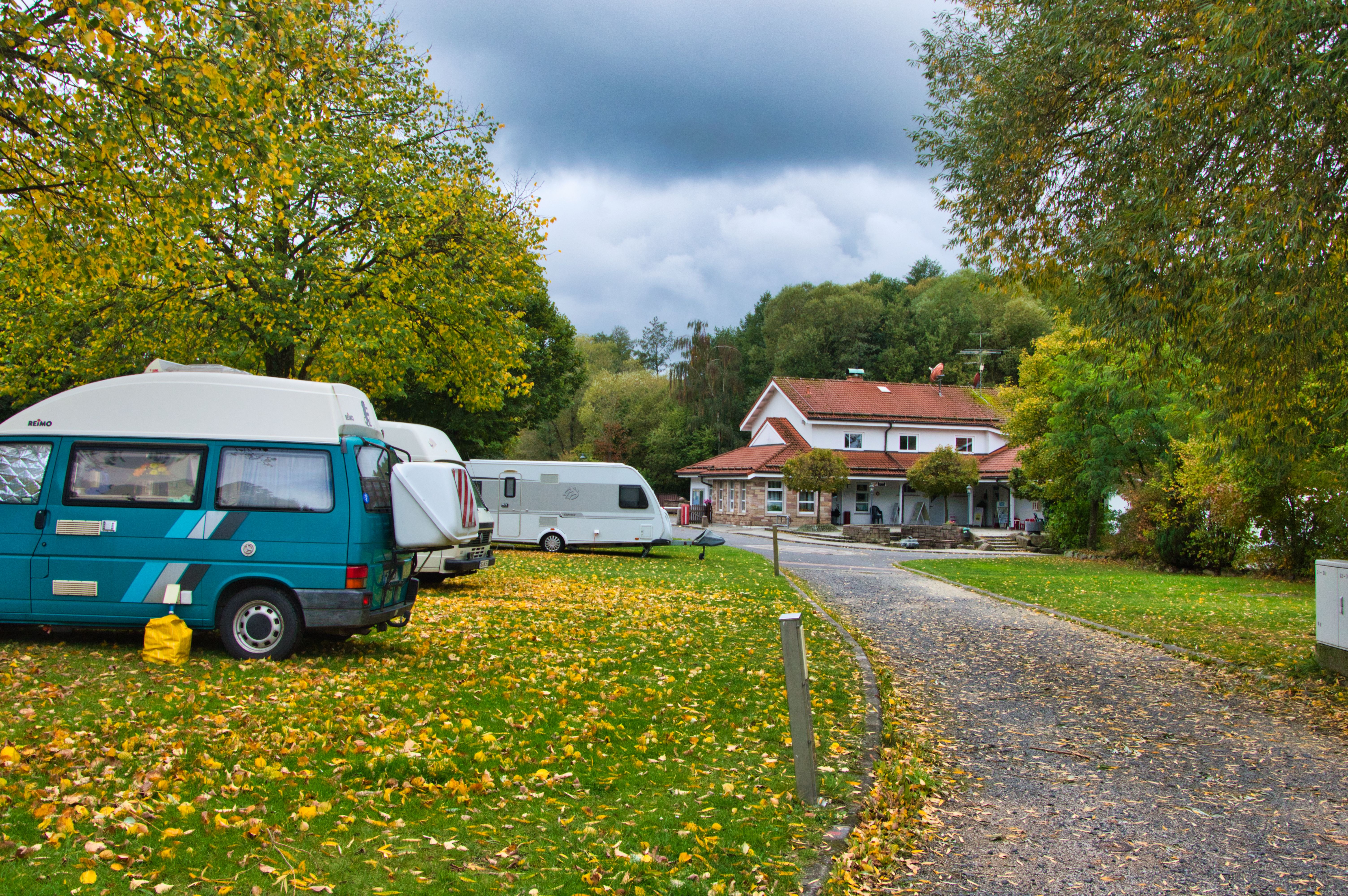 Wohnmobile auf einem Kiesweg am Campingplatz, im Hintergrund Gebäude mit rotem Dach und viel Grün.