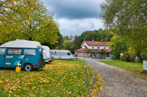 Wohnmobile auf einem Kiesweg am Campingplatz, im Hintergrund Gebäude mit rotem Dach und viel Grün.