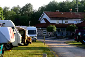 Wohnmobile und Campingbusse stehen auf einem Grasplatz am Weg, im Hintergrund ein Gebäude mit rotem Ziegeldach und Bäumen.