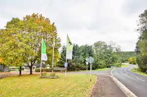 Eingang eines Campingplatzes mit zwei grünen Flaggen, Schild am Weg und Straße am Rand mit Bäumen und Hecken.