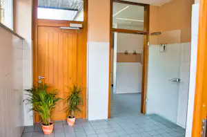 Holztür und offene Tür zum Sanitärraum mit Fliesenboden, Waschbeckenarmaturen und zwei Haken an der Wand.