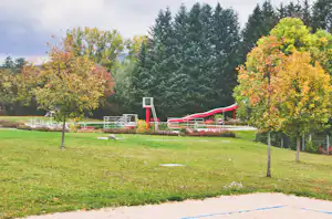 Rote Rutsche auf einem Spielplatz im Grünen, umgeben von Bäumen, Rasen und einem Sandbereich.