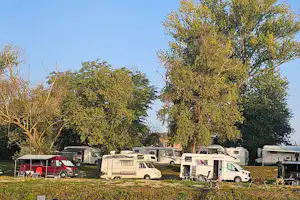 Reihe von Wohnmobilen auf einem Campingplatz zwischen Bäumen, mit Vorzelten und Campingstühlen am Stellplatz.