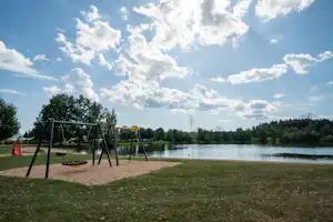 Schaukel im Spielplatz am See, Rasen und Sandfläche am Ufer, Bäume und Strommasten im Hintergrund.