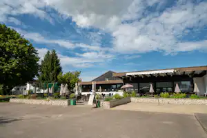 Terrasse am Campingplatz mit Tischen und Stühlen unter weißen Sonnenschirmen vor einem modernen Gebäude, umgeben von Bäumen.