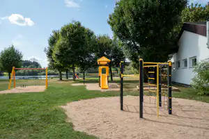 Spielplatz am See mit gelbem Klettergerüst, Sandboden, Schaukel und Bäumen neben dem weißen Gebäude.