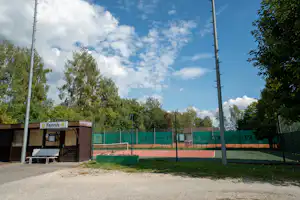 Tennisplatz auf dem Campinggelände mit Netz, Zaun und Umkleidehäuschen, umgeben von Bäumen und blauem Himmel.