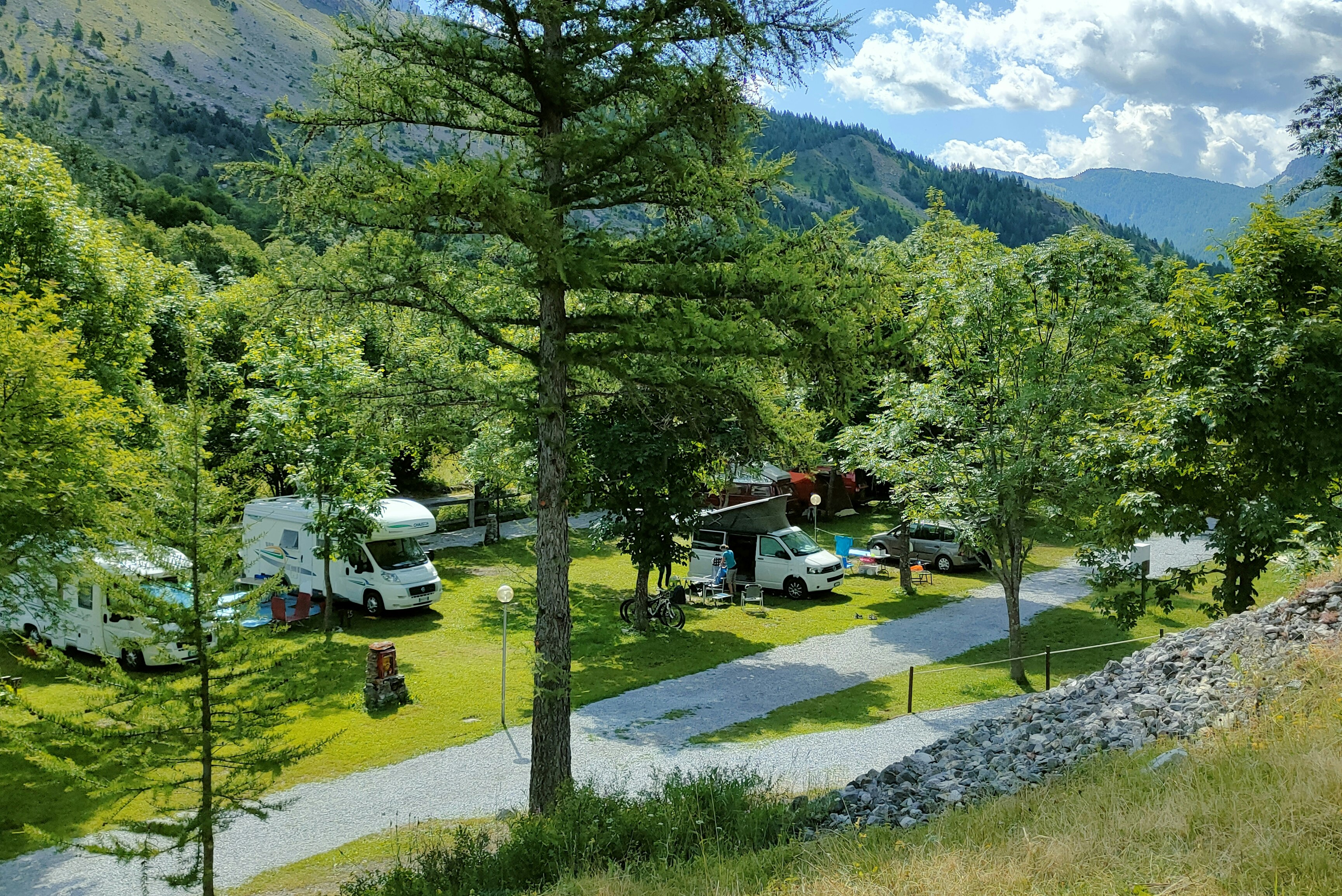 Campingplatz im Grünen mit Wohnmobilen und Wohnwagen, Kiesweg und Bäumen vor einem grünen Tal und Bergen.