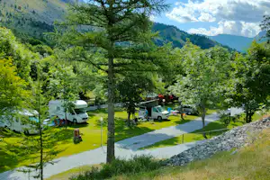 Campingplatz im Grünen mit Wohnmobilen und Wohnwagen, Kiesweg und Bäumen vor einem grünen Tal und Bergen.