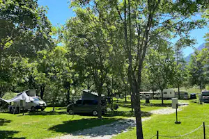 Campingplatz mit Wohnmobilen und Zelten zwischen hohen Bäumen, grüner Wiese und Blick auf Berge im Hintergrund.