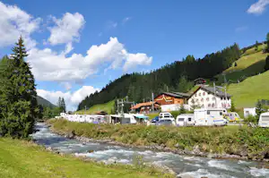 Wohnmobilstellplatz am Fluss mit vielen Campern, Wohngebäuden und grünen Hängen im Tal unter blauem Himmel.