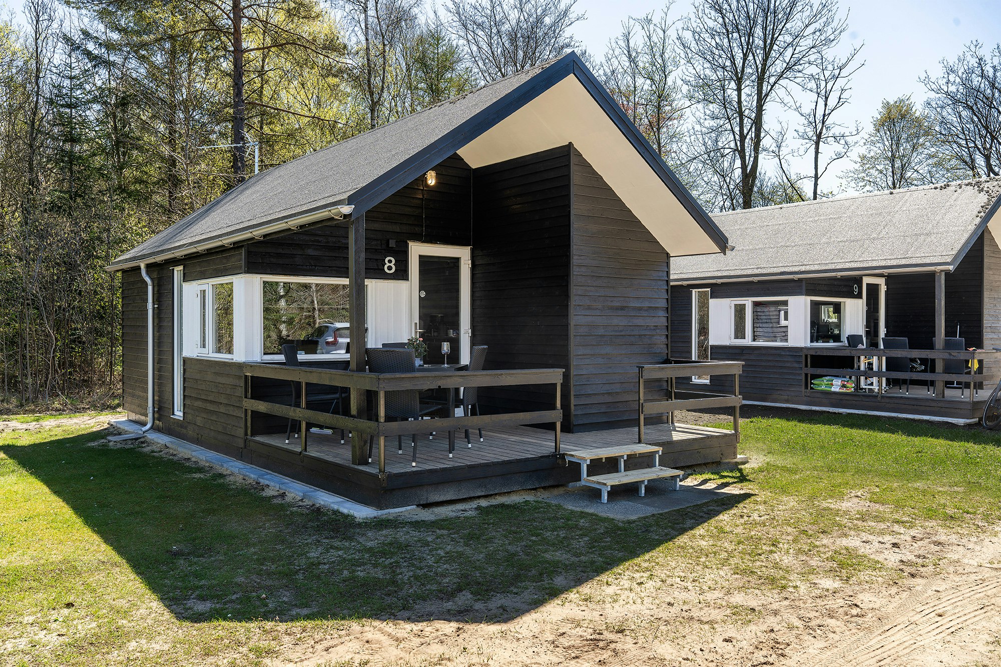 Holz-Campinghaus mit überdachter Terrasse, Sitzbank und Tisch, daneben weitere Unterkunft mit Veranda im Grünen am Waldrand.