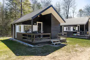 Holz-Campinghaus mit überdachter Terrasse, Sitzbank und Tisch, daneben weitere Unterkunft mit Veranda im Grünen am Waldrand.