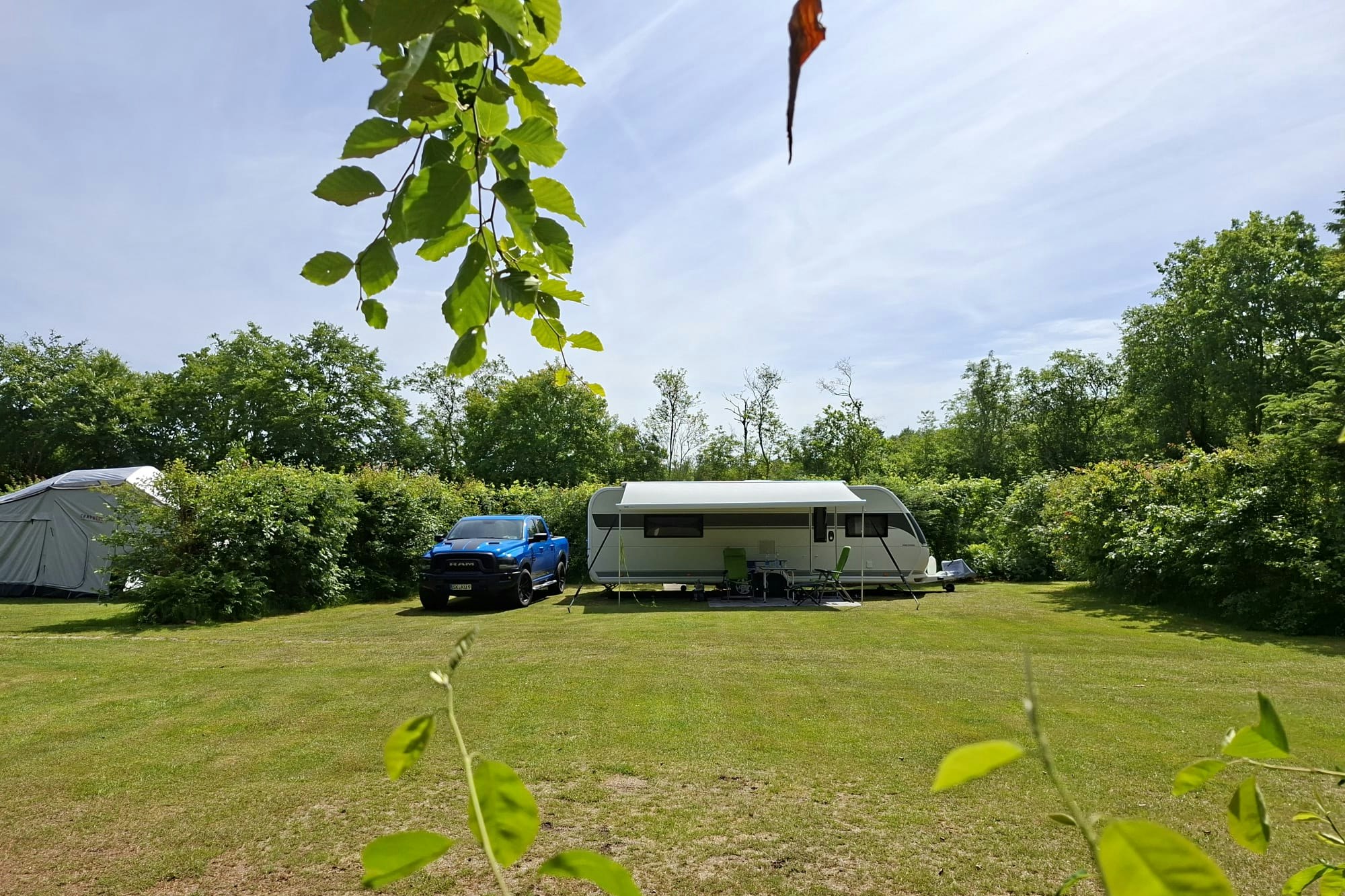 Wohnwagen und Auto auf einem gepflegten Campingplatz, umgeben von Bäumen und Sträuchern, mit Rasenfläche im Vordergrund.