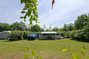 Wohnwagen und Auto auf einem gepflegten Campingplatz, umgeben von Bäumen und Sträuchern, mit Rasenfläche im Vordergrund.