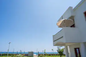 Weißes Ferienhaus mit Balkon und roten Fenstern, davor Garten mit Büschen und Blick auf das Meer am Horizont.
