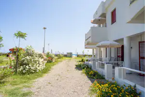 Weiße Ferienhaus-Reihe mit Terrasse, Blumenbeeten und Kiesweg am Campingplatz, im Hintergrund Zaun und Laternen.