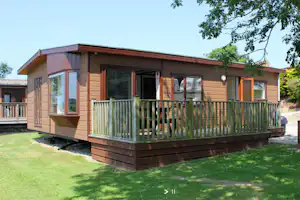 Modernes Mobilheim mit Veranda und Holzgeländer auf dem Campingplatz, umgeben von Rasen und Bäumen.