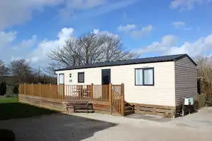 Mobilheim mit Holzterrasse, Sitzbank und Schotterweg vor dem Haus, umgeben von Rasen und Bäumen.