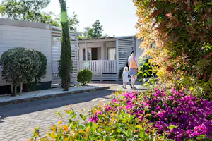 Gepflasterter Weg am Campingplatz mit Blumenbeeten, Hecken und einer Terrasse mit Holzgeländer neben einem Wohngebäude.