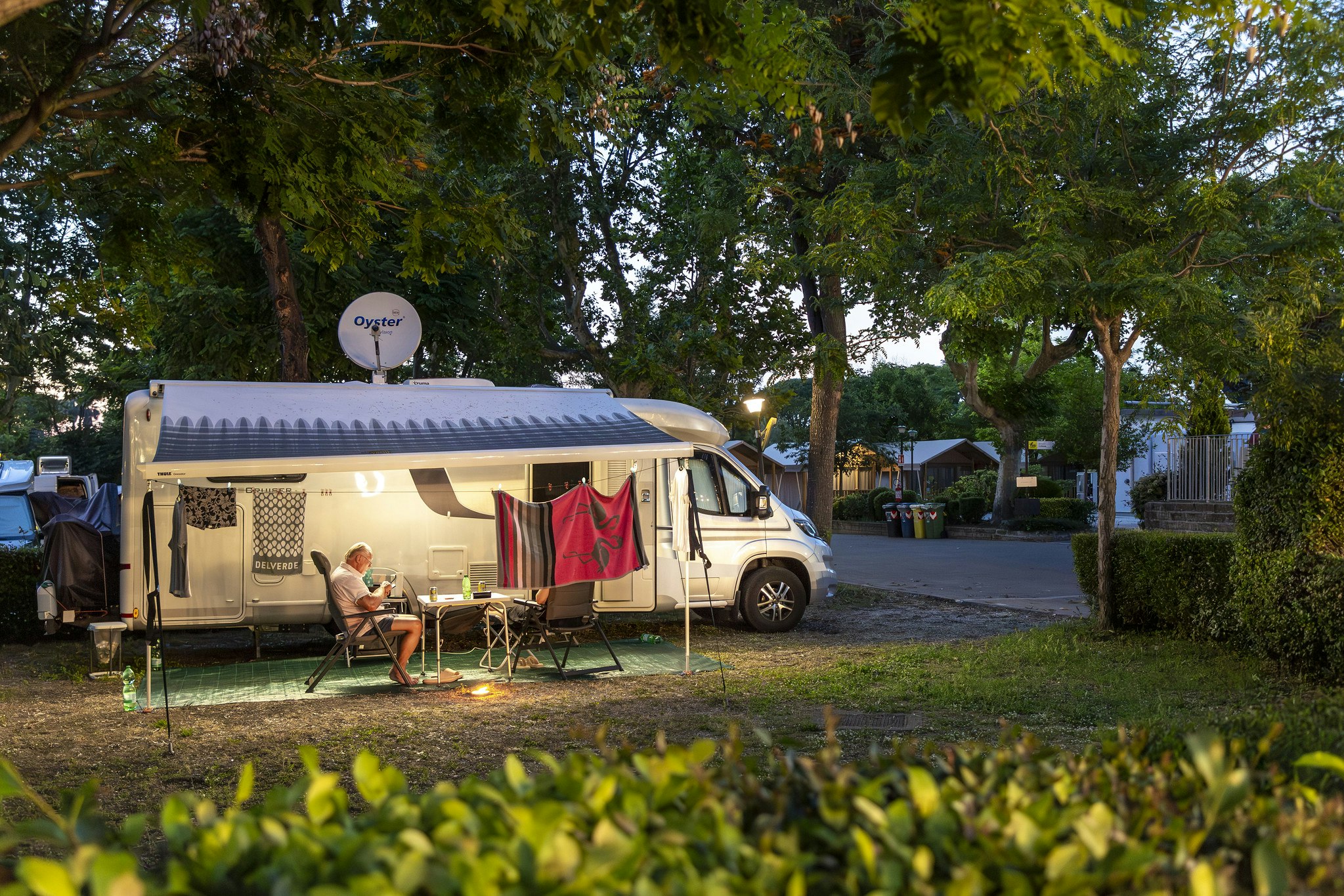 Wohnwagen mit Vorzelt und kleiner Terrasse, Campingstuhl und Tisch davor, im Hintergrund Bäume und weitere Stellplaetze.