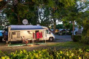 Wohnwagen mit Vorzelt und kleiner Terrasse, Campingstuhl und Tisch davor, im Hintergrund Bäume und weitere Stellplaetze.