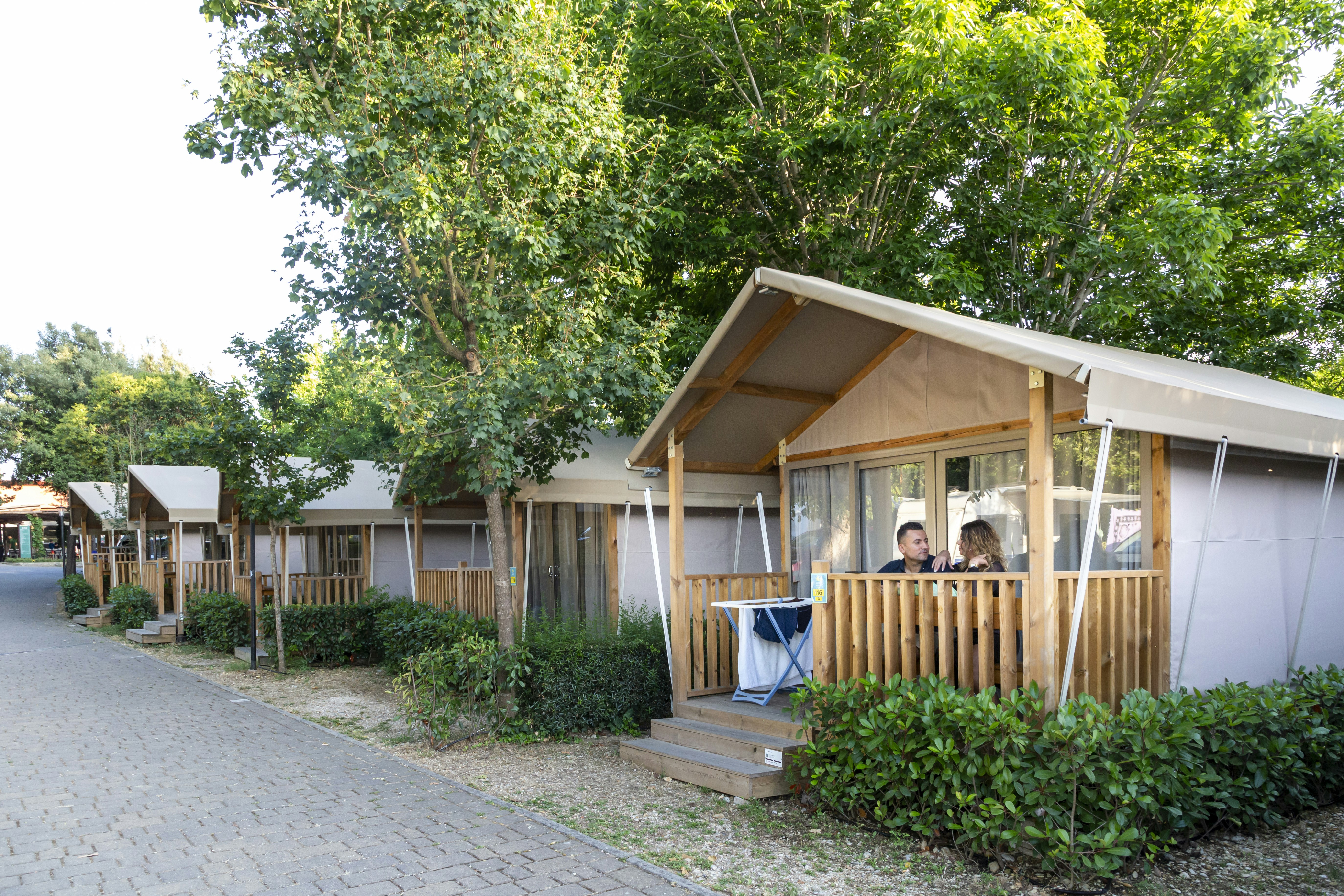 Holz- und Zeltbungalow mit Veranda und Sitzbank, am Wegesrand zwischen Büschen und Bäumen auf dem Campingplatz.